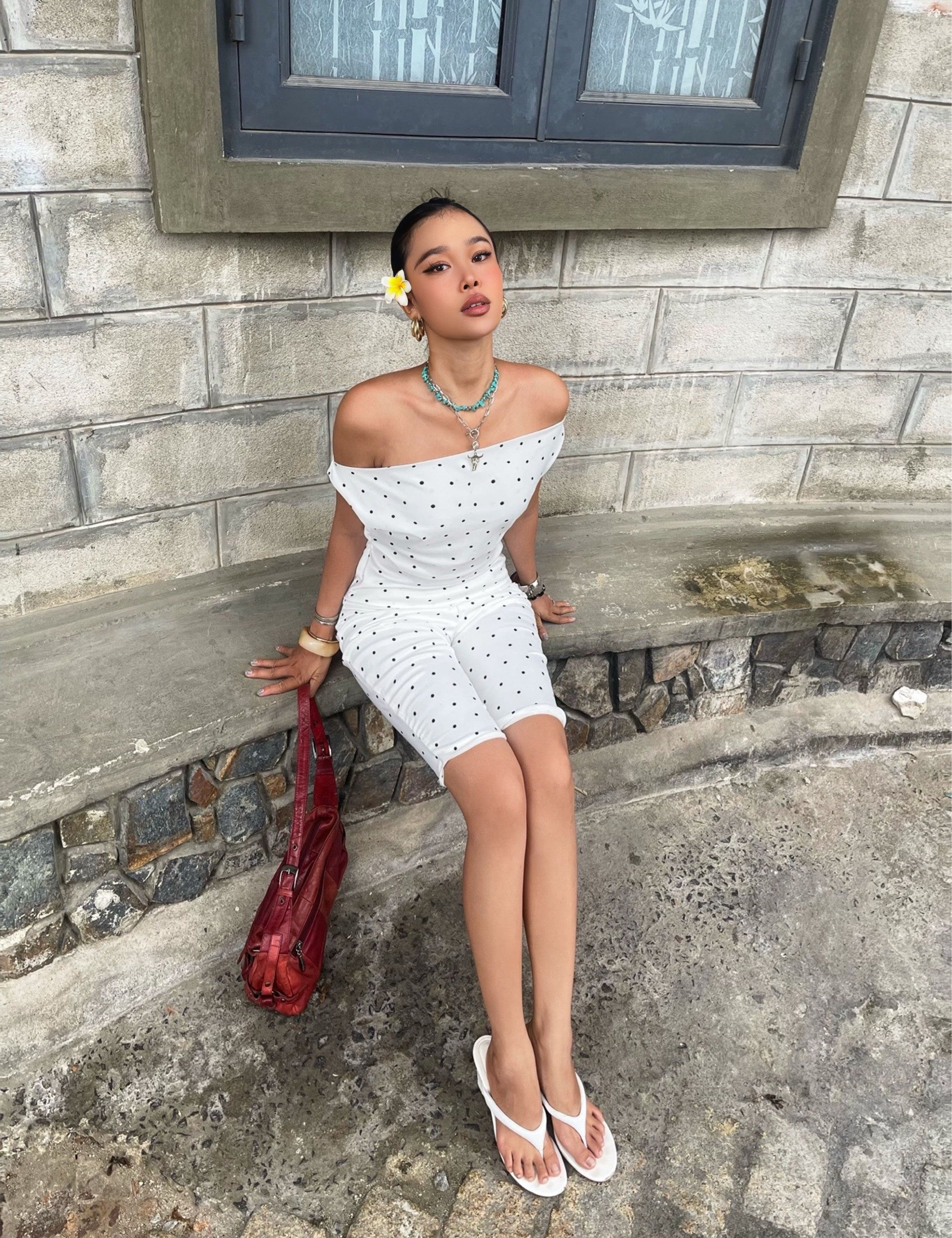 Woman wearing Clara Polka Dot Satin Set, sitting outside on a stone bench near a window, white sandals and red bag.
