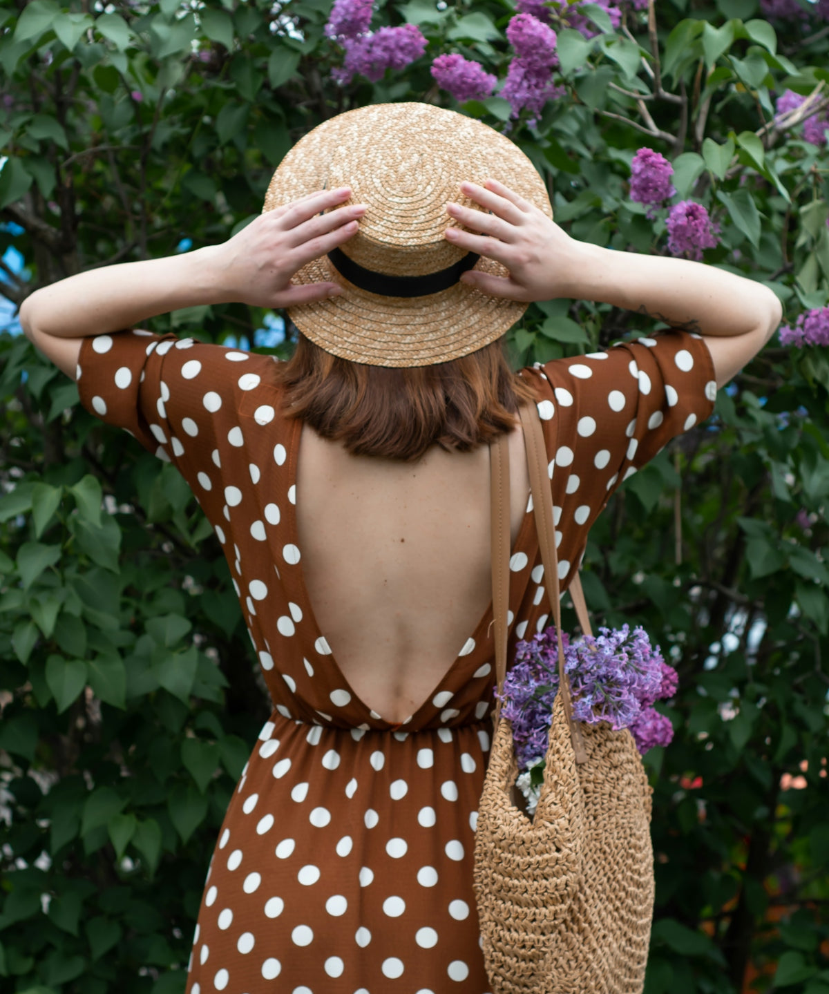 woman in white and black polka dot dress wearing brown straw hat standing near purple flowers
