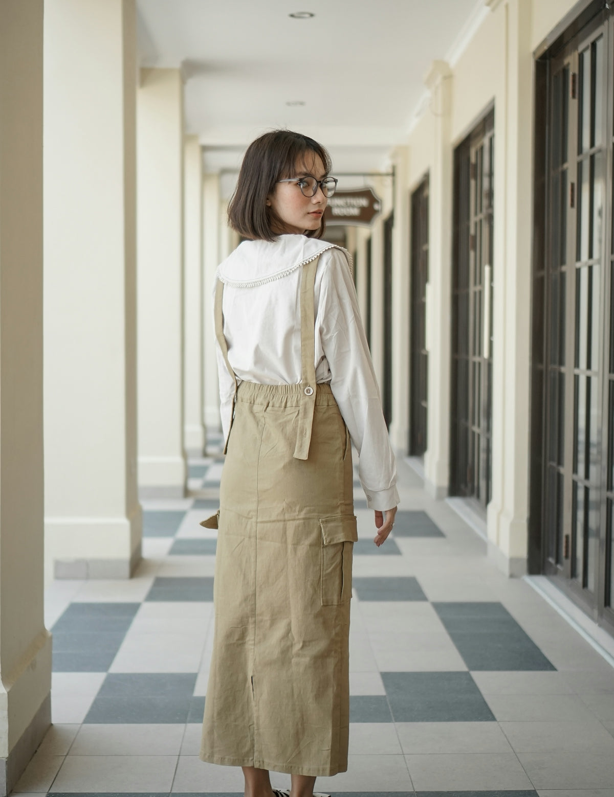 A woman standing in a hallway wearing a white shirt and khaki skirt