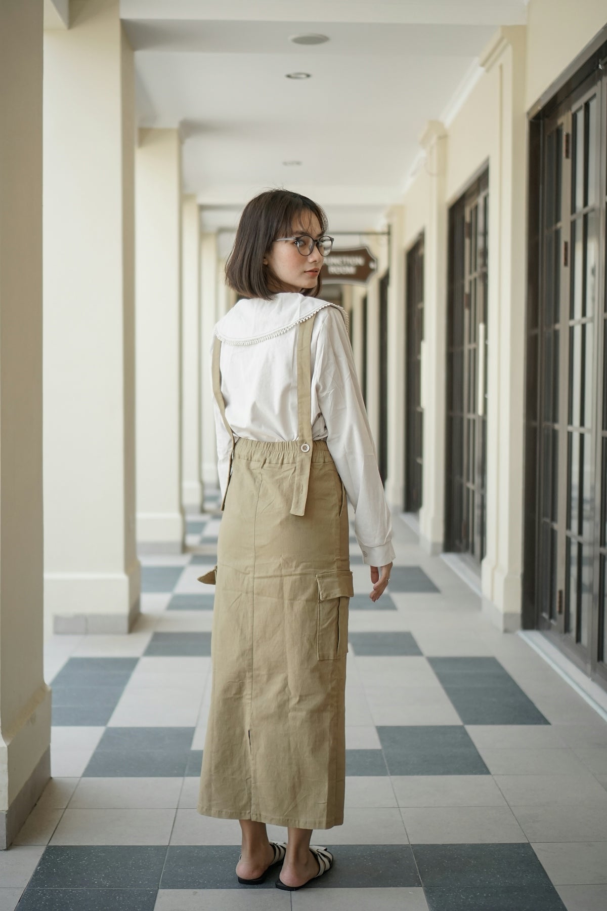 A woman standing in a hallway wearing a white shirt and khaki skirt