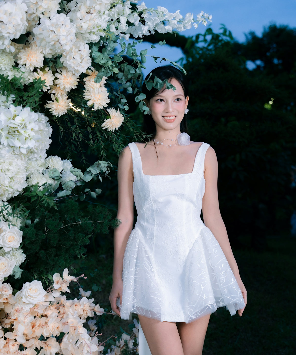A woman in a white dress stands near floral decorations.