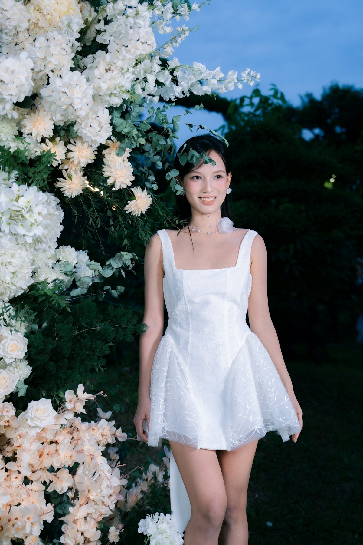 A woman in a white dress stands near floral decorations.